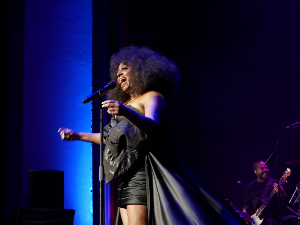 Ledisi performs onstage at the Paramount Theatre in Denver, smiling and singing into a microphone while wearing a dramatic black outfit with a long cape. Blue stage lighting illuminates the background, and a band member plays guitar behind her.