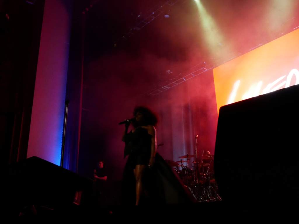 Ledisi stands onstage in low, moody lighting, holding a microphone as red and purple fog surrounds her. A large LED screen behind her displays her name, and the band plays in the background.