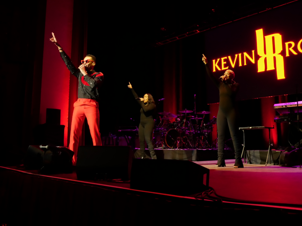 Kevin Ross performs with two backup singers at the Paramount Theatre, all raising their arms in unison. Bright red lighting illuminates the stage and a large screen behind them displays his name in bold lettering.