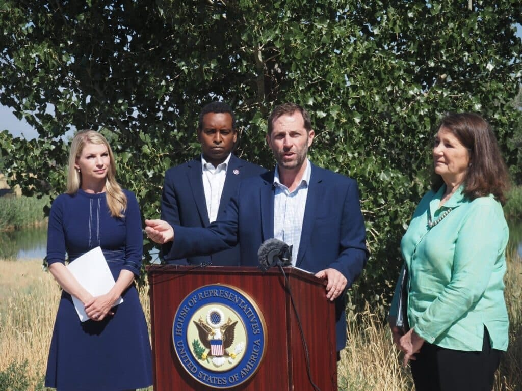 Four Colorado lawmakers stand outdoors behind a podium with the U.S. House of Representatives seal. One man speaks into microphones while the others—two women and another man—stand beside him, listening. Trees and tall grass appear in the background.
