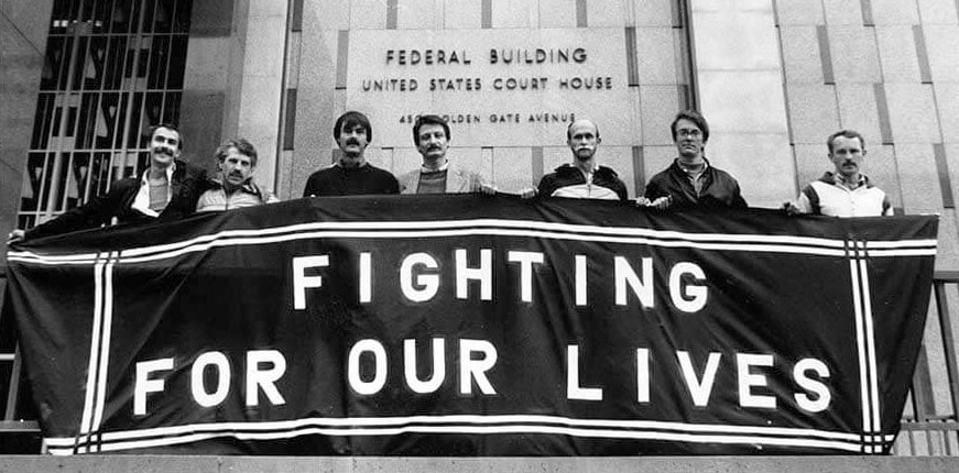 A black-and-white photograph of seven men standing in front of the San Francisco Federal Building, holding a large banner that reads “FIGHTING FOR OUR LIVES.” The men appear solemn and determined, representing early HIV/AIDS activism during the height of the crisis.