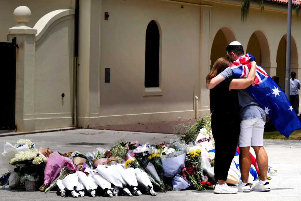 Two people embrace at a makeshift memorial of flowers outside a building in Sydney, with one draped in an Australian flag, honoring victims of the Bondi Beach shooting.