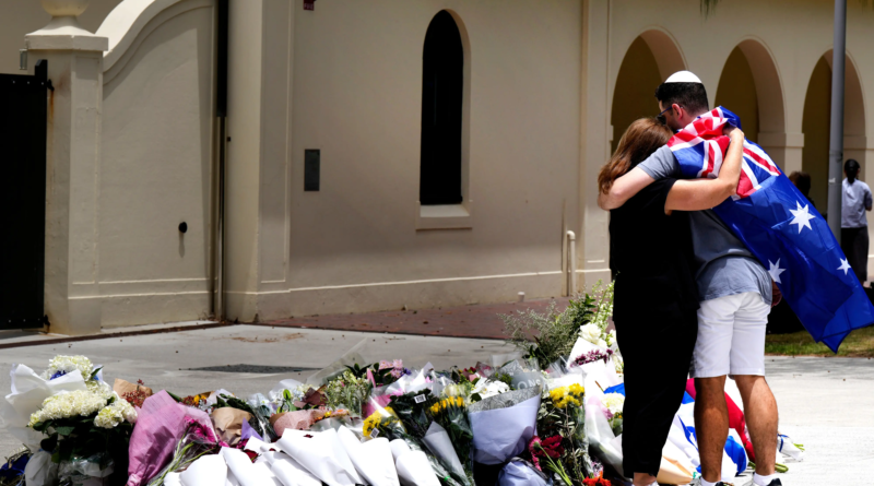 Two people embrace at a makeshift memorial of flowers outside a building in Sydney, with one draped in an Australian flag, honoring victims of the Bondi Beach shooting.
