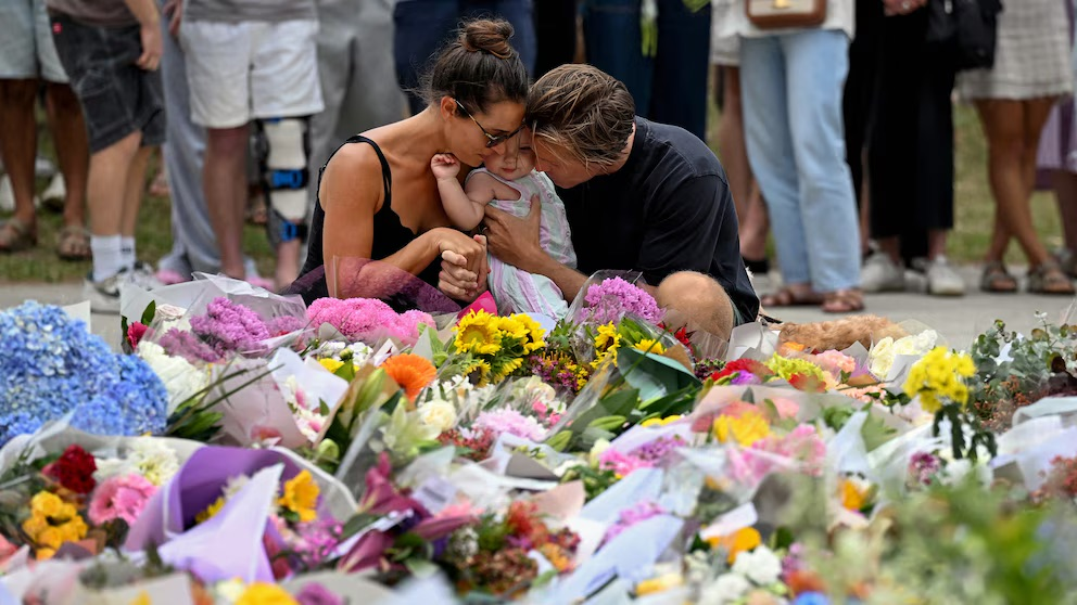 A couple holds a baby while kneeling among bouquets of flowers at a memorial honoring victims of the Bondi Beach shooting, as mourners gather in the background.