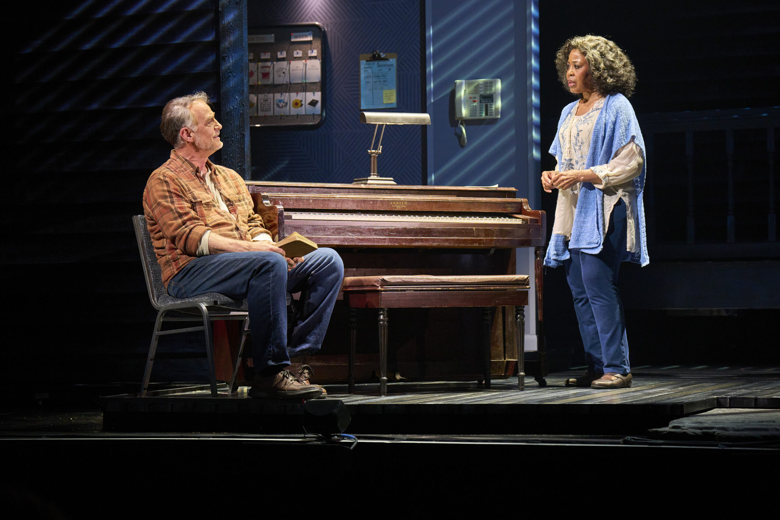 An older couple stands and sits beside a piano onstage during a performance of The Notebook musical, sharing a quiet, emotional moment under soft stage lighting.