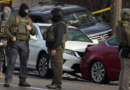 Armed law enforcement officers wearing tactical gear stand in the street near several damaged vehicles and yellow police tape at the scene of a shooting in a residential area of Minneapolis during a federal immigration enforcement operation.