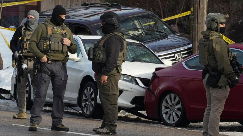 Armed law enforcement officers wearing tactical gear stand in the street near several damaged vehicles and yellow police tape at the scene of a shooting in a residential area of Minneapolis during a federal immigration enforcement operation.
