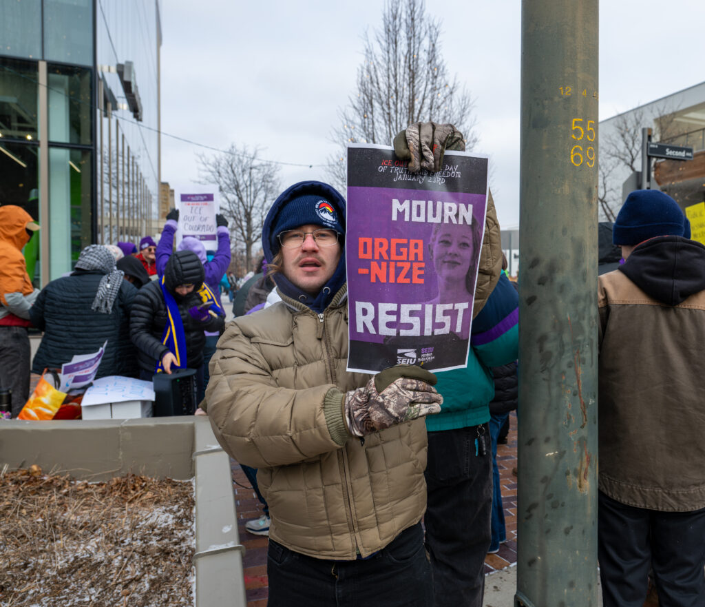 Crowd of demonstrators bundled in winter clothing gather on a sidewalk in Denver’s Cherry Creek neighborhood, holding signs criticizing ICE and corporate involvement in immigration enforcement.