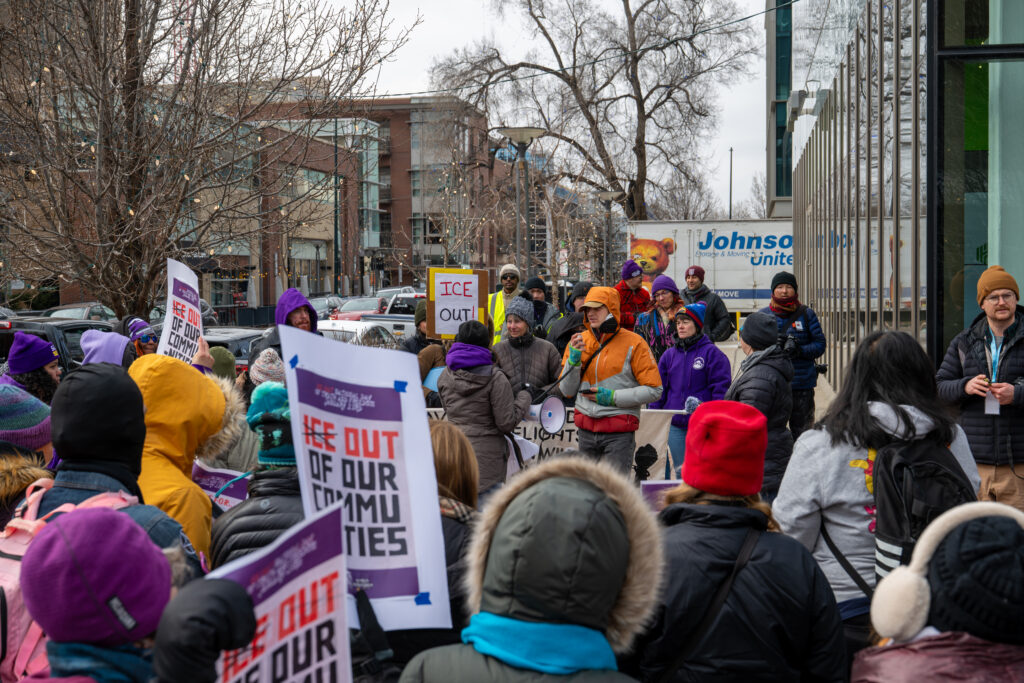 A speaker addresses a crowd through a megaphone while protesters hold signs calling for ICE out of Colorado during a labor- and community-led rally outside Palantir Technologies in Denver.