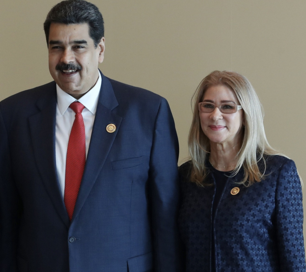 A man and woman in formal attire stand side by side indoors, both wearing dark suits and official pins, posing for a photograph against a plain background during a diplomatic or governmental event.