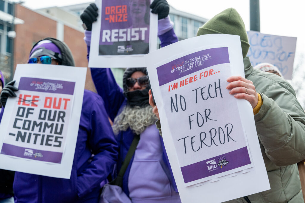 Group of protesters in winter coats hold signs reading “ICE out of our communities” and “No tech for terror” during a rally outside Palantir Technologies’ Denver office.