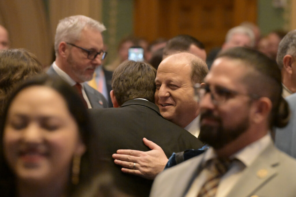 Gov. Jared Polis embraces an attendee in a crowded room, smiling as others stand nearby in the foreground and background.