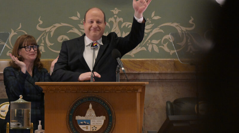 Gov. Jared Polis waves from the podium during the State of the State while House Speaker Julie McCluskie applauds beside him.