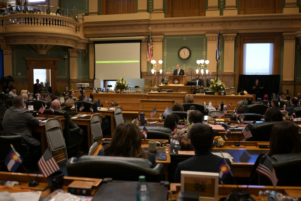 Wide view of the Colorado House chamber during the State of the State, with lawmakers seated at desks facing the dais as the governor speaks at a lectern beneath U.S. and Colorado flags.
