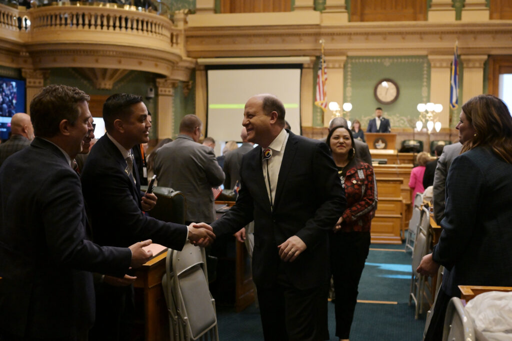 Gov. Jared Polis smiles and shakes hands with lawmakers on the House floor as people mingle in the chamber after the address.