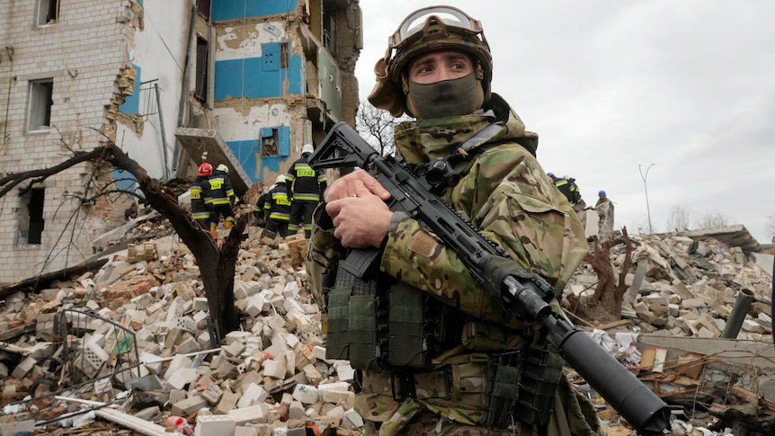 Armed soldier in camouflage and protective gear stands in front of a heavily damaged residential building, with rubble and emergency responders visible in the background at a conflict site.