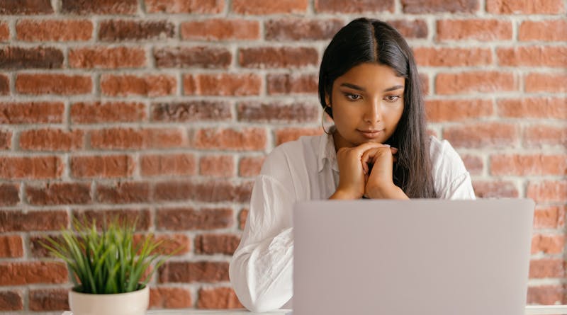 Young woman deeply focused on her laptop studying indoors against a brick wall backdrop.