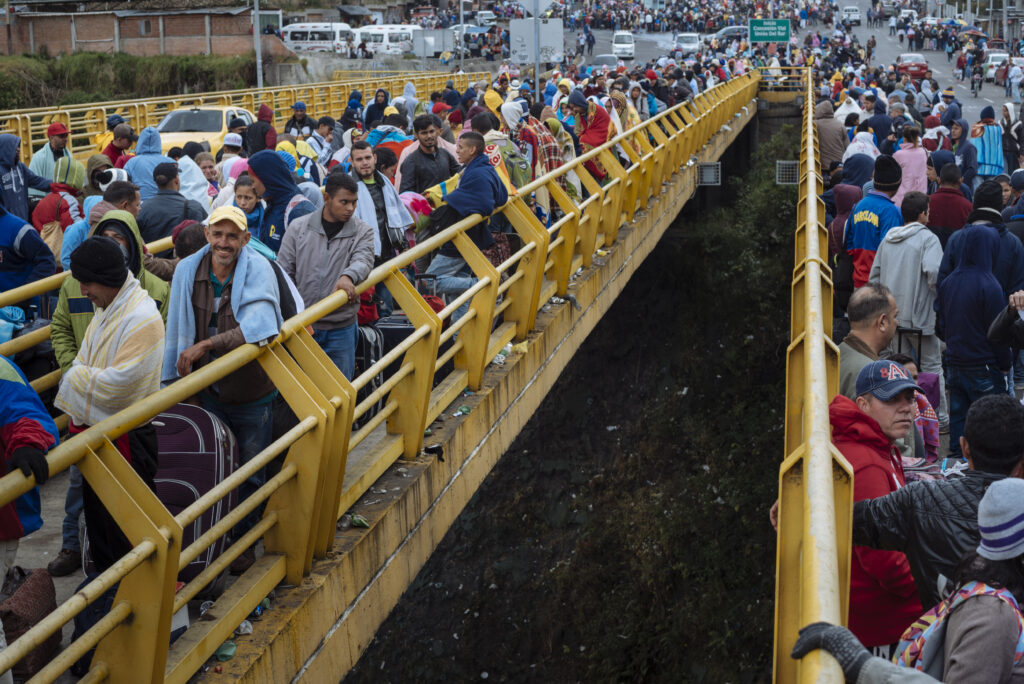 Large crowds of people carrying bags and belongings cross a yellow bridge, many wearing jackets and blankets, as part of a mass migration out of Venezuela amid ongoing economic and political instability.