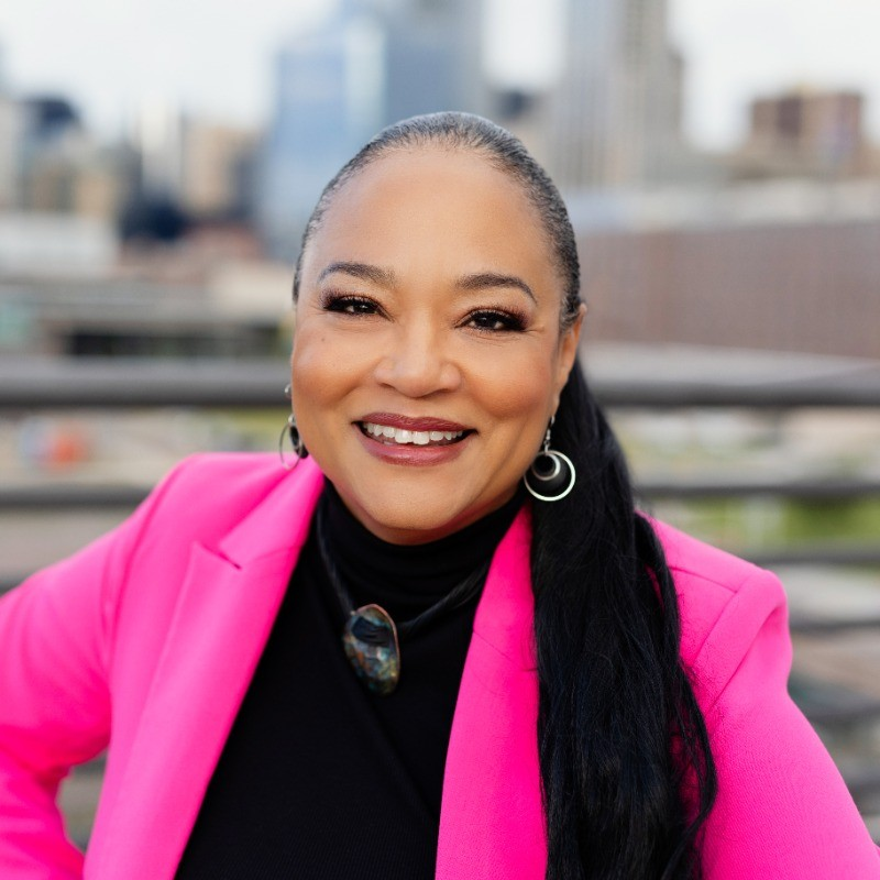 Smiling woman wearing a bright pink blazer over a black top, with long dark hair pulled back into a low ponytail, standing outdoors on a rooftop with a blurred city skyline in the background.