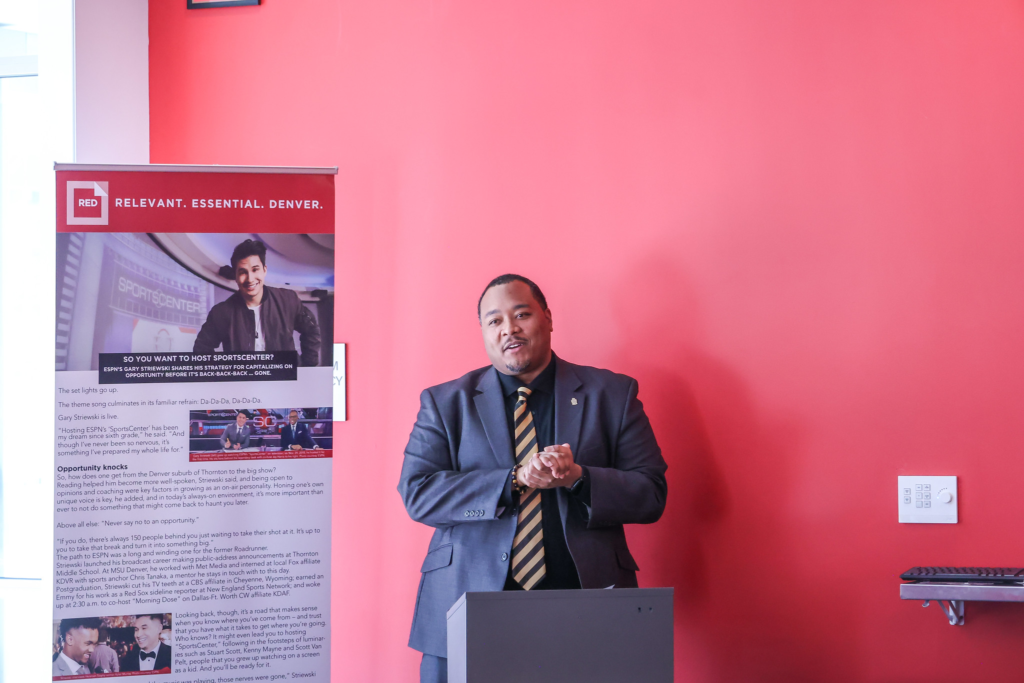 A man in a dark suit and gold-striped tie speaks at a podium against a bright red wall at MSU Denver, with a banner promoting student success visible beside him.