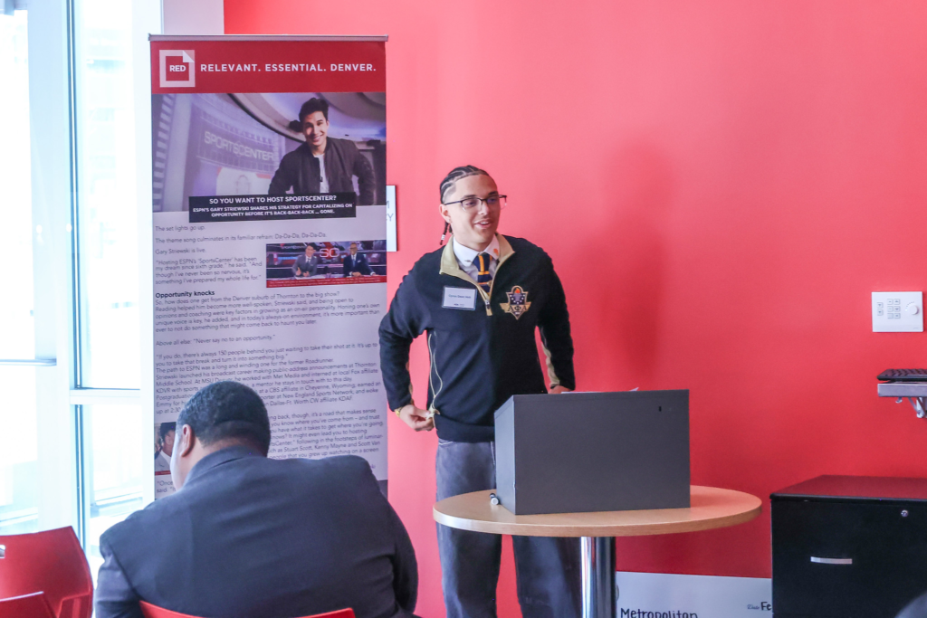 A student wearing glasses and a black-and-gold Alpha Phi Alpha jacket speaks at a podium during the campus event, with attendees seated in front of him and a red wall backdrop behind.