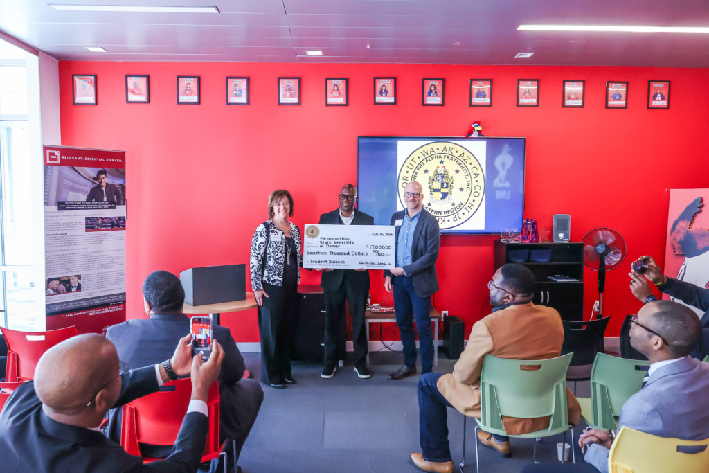 Three officials stand in front of a red wall at Metropolitan State University of Denver holding a ceremonial $17,000 check for student success initiatives, while attendees seated in the foreground photograph the presentation. A screen behind them displays the Alpha Phi Alpha Western Region emblem.