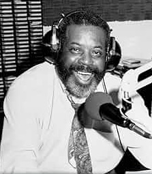 Black-and-white photo of Dr. Daddio smiling in a radio studio, wearing headphones and speaking into a large broadcast microphone, seated in front of shelves lined with CDs.
