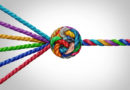 Close-up of multiple brightly colored braided ropes—red, blue, green, purple, yellow, and teal—converging from opposite sides and tied together in a tight central knot against a light gray background.