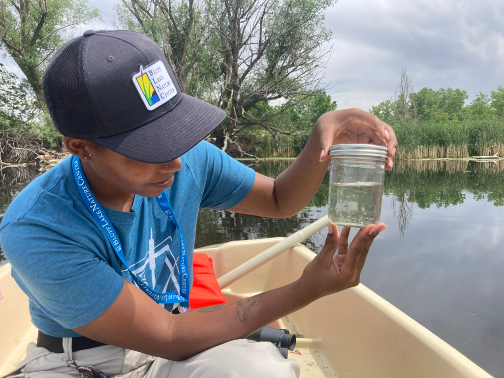 A Bluff Lake Nature Center staff member wearing a Bluff Lake cap and lanyard examines a clear jar of pond water while seated in a small boat, demonstrating hands-on environmental education and water quality exploration on the lake.
