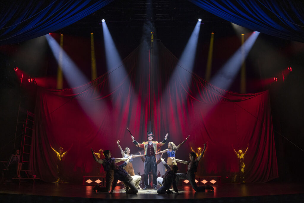 A ringmaster stands center stage beneath dramatic spotlights, arms raised as the circus ensemble kneels and extends their arms around him against a sweeping red velvet backdrop shaped like a tent.