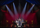 A ringmaster stands center stage beneath dramatic spotlights, arms raised as the circus ensemble kneels and extends their arms around him against a sweeping red velvet backdrop shaped like a tent.