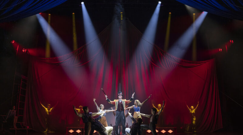 A ringmaster stands center stage beneath dramatic spotlights, arms raised as the circus ensemble kneels and extends their arms around him against a sweeping red velvet backdrop shaped like a tent.