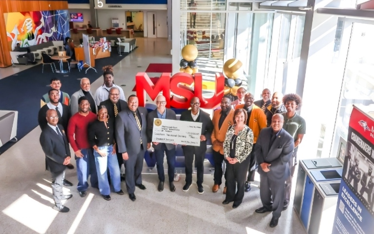 Group photo inside MSU Denver’s atrium shows fraternity members, university leaders and students gathered around a large ceremonial $17,000 check in front of oversized red “MSU” letters and black-and-gold balloon décor.