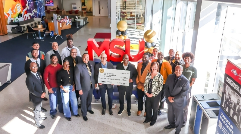 Group photo inside MSU Denver’s atrium shows fraternity members, university leaders and students gathered around a large ceremonial $17,000 check in front of oversized red “MSU” letters and black-and-gold balloon décor.