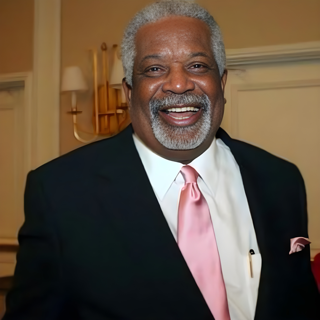 Alt text: Dr. Daddio, an older Black man with silver hair and a neatly groomed gray mustache and goatee, smiles warmly while wearing a black suit, white dress shirt and pink tie, standing indoors against a softly lit neutral background.