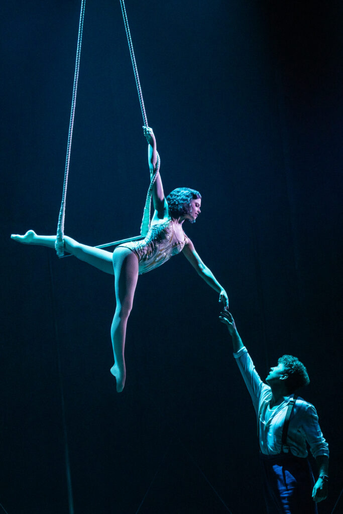 An aerialist suspended from two ropes reaches down toward a man below her, illuminated by cool blue lighting in an intimate circus performance scene from Water for Elephants.