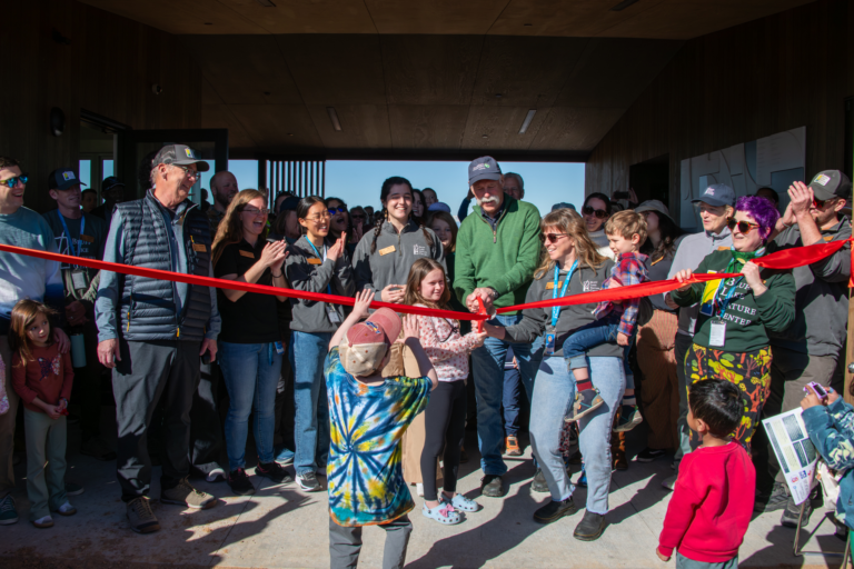 Lead donor David Smith, Executive Director Rachel Hutchens and camper Lily Prutch cut a bright red ribbon at the entrance of Bluff Lake Nature Center during the Feb. 14, 2026 grand opening celebration, surrounded by staff, families and community members applauding inside the new net-zero facility lobby.