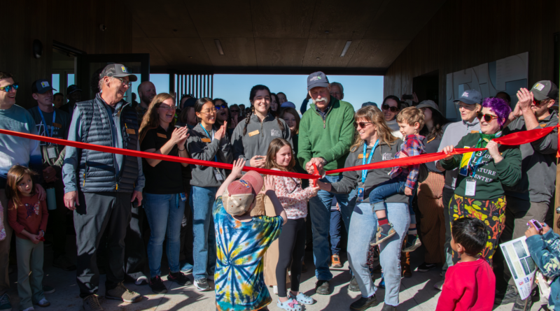Lead donor David Smith, Executive Director Rachel Hutchens and camper Lily Prutch cut a bright red ribbon at the entrance of Bluff Lake Nature Center during the Feb. 14, 2026 grand opening celebration, surrounded by staff, families and community members applauding inside the new net-zero facility lobby.