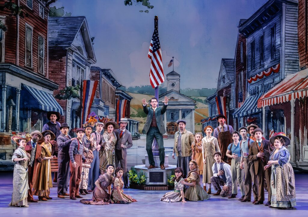 Full cast of The Music Man gathered in a painted Midwestern town square set, with Harold Hill standing elevated beneath an American flag as townspeople in period costumes look on.