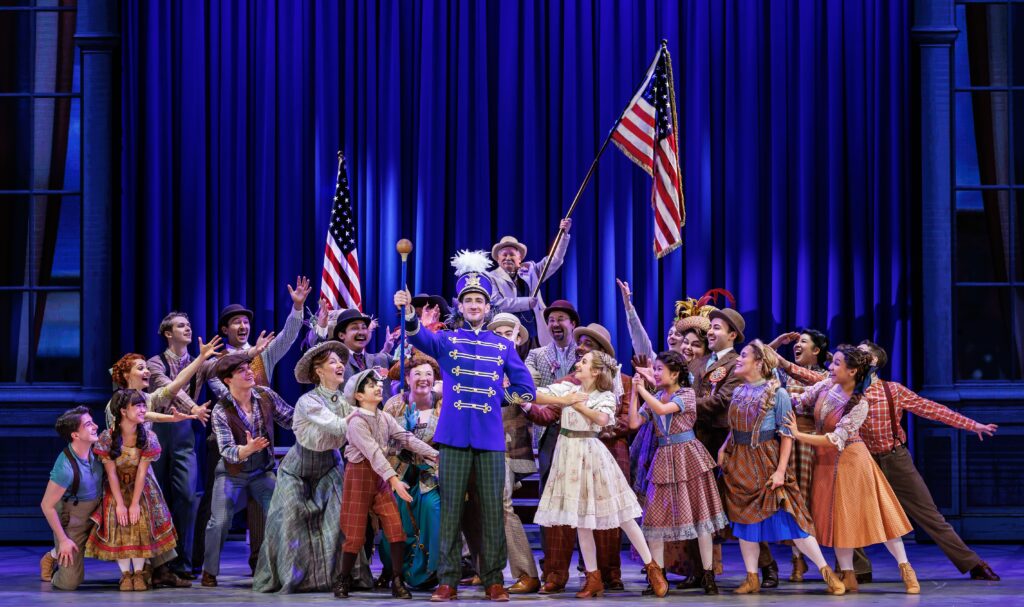 Harold Hill in a bright blue band uniform stands center stage surrounded by an enthusiastic ensemble in colorful early 1900s attire, celebrating beneath American flags and rich blue lighting.