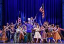 Harold Hill in a bright blue band uniform stands center stage surrounded by an enthusiastic ensemble in colorful early 1900s attire, celebrating beneath American flags and rich blue lighting.