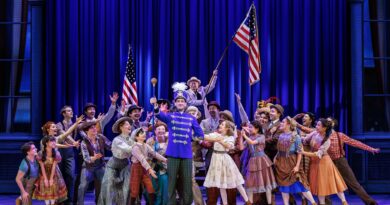 Harold Hill in a bright blue band uniform stands center stage surrounded by an enthusiastic ensemble in colorful early 1900s attire, celebrating beneath American flags and rich blue lighting.