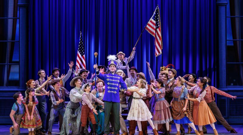 Harold Hill in a bright blue band uniform stands center stage surrounded by an enthusiastic ensemble in colorful early 1900s attire, celebrating beneath American flags and rich blue lighting.