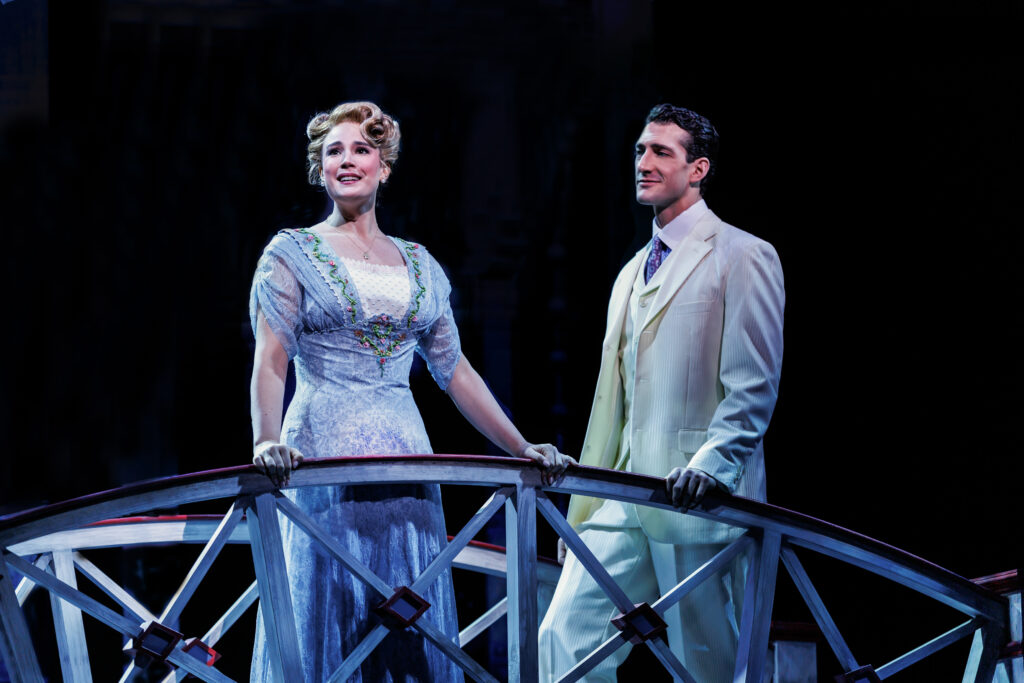 Marian Paroo and Harold Hill stand together on a footbridge under soft stage lighting, wearing elegant Edwardian costumes during a romantic moment in The Music Man.