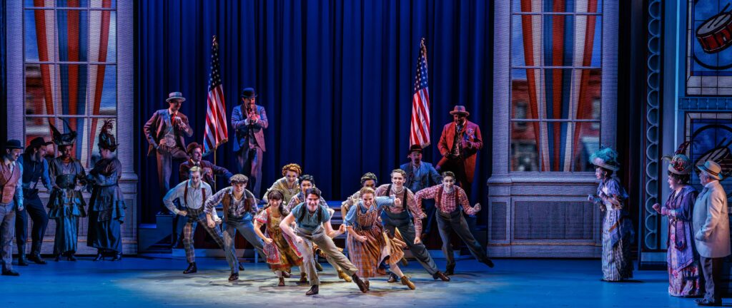 Ensemble cast members perform energetic choreography in front of tall American flags and stylized storefront windows, dressed in period costumes reflecting an early 20th-century Midwestern town.