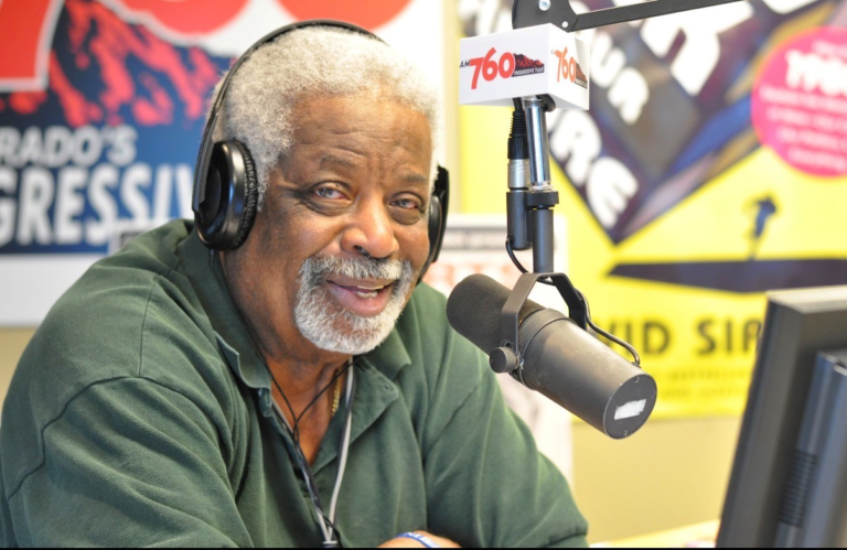 Dr. Daddio sits in a radio studio wearing headphones and speaking into a broadcast microphone, smiling toward the camera in front of colorful station signage and equipment.