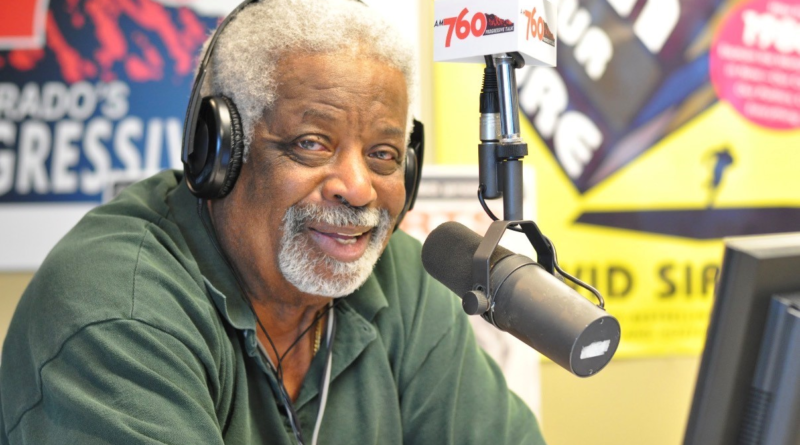 Dr. Daddio sits in a radio studio wearing headphones and speaking into a broadcast microphone, smiling toward the camera in front of colorful station signage and equipment.