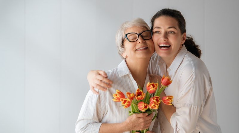 An older woman and a younger woman embrace while holding a bouquet of tulips, smiling together indoors.