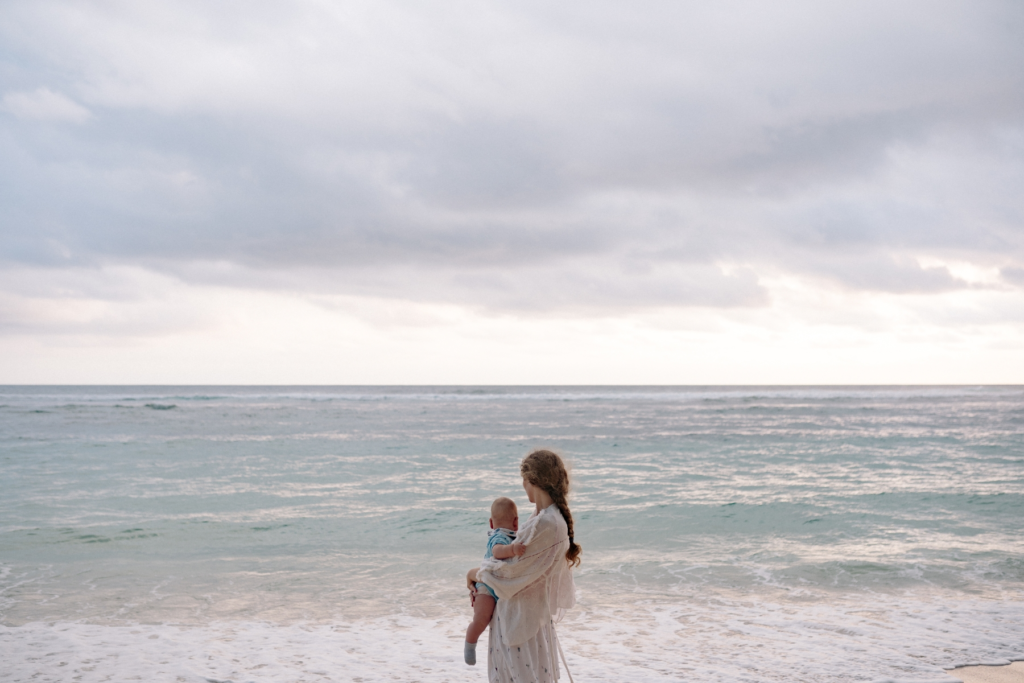 A woman stands on a beach holding a baby while looking out at the ocean under a cloudy sky.