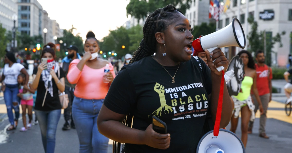 Protester holding a megaphone leads a march, wearing a shirt that reads “Immigration is a Black Issue,” with a crowd gathered behind her in a city street.